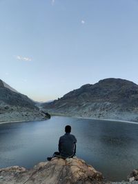 Rear view of man sitting on rock by mountain against sky