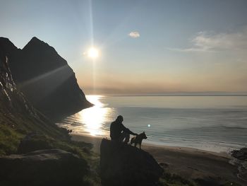 Man with dog sitting on rocks by sea against sky during sunset