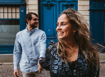 Happy young woman in stylish outfit followed by smiling boyfriend walking on city street with old building in background during romantic holidays in france