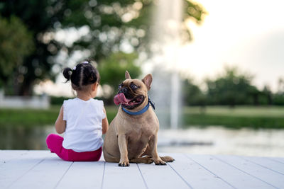 Rear view of girl sitting by dog looking away against fountain