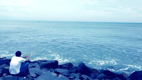 Rear view of man sitting by sea against sky