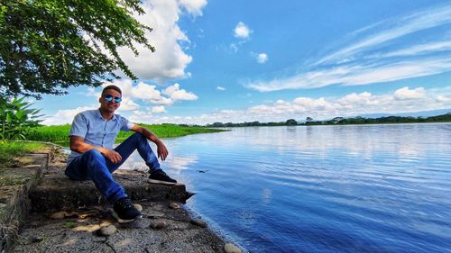 Young man sitting on rock by lake against sky