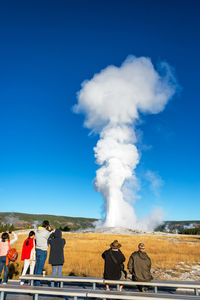 People looking at geyser against blue sky