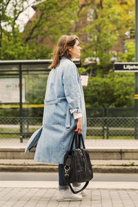 Full length of young woman holding disposable coffee cup while waiting at tram station