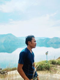 Side view of young man looking away while standing by lake against mountains and sky