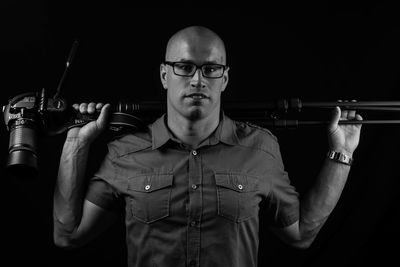 Portrait of bald young man with camera on tripod standing against black background