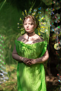 Young woman in mirrored mask and neon fabric standing in forest  front of the scenery from a cd case