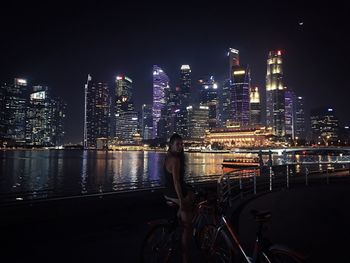 Woman with bicycle against illuminated buildings in city at night