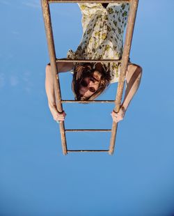 Low angle portrait of young man climbing on ladder against clear blue sky