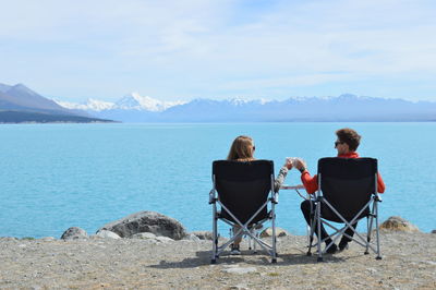 Rear view of couple sitting on chair by lake against sky