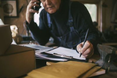 Cropped image of gay man writing on paper while talking over smart phone at garage
