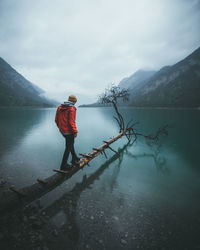 Rear view of man standing on tree trunk over lake against mountains during winter