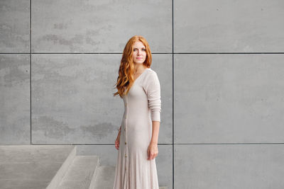 Positive young female with long red hair wearing casual dress and looking at camera while standing against concrete wall