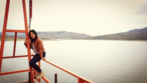 Young woman sitting on railing by lake against sky