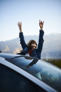 Young woman with arms raised and tousled hair sitting in car