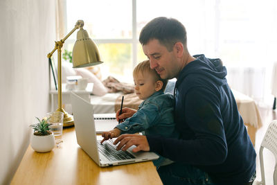 Side view of man with boy in lap using laptop at home