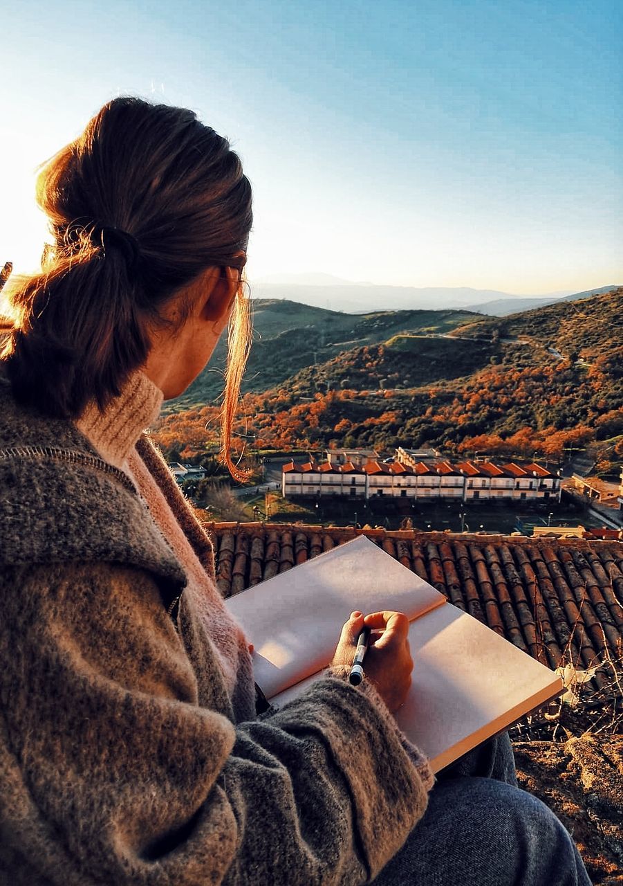 one person, young adult, mountain, leisure activity, sitting, real people, lifestyles, nature, scenics - nature, sky, architecture, young women, adult, three quarter length, landscape, casual clothing, women, environment, building exterior, outdoors, looking at view, cityscape, hairstyle