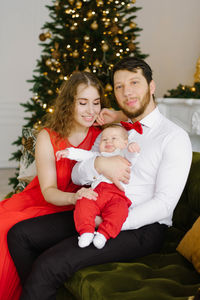 A young happy family with a child is sitting on a chair near a decorated christmas tree person