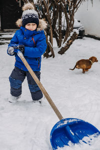 Little boy with a shovel in hand remove snow in backyard, snow removal. kid in blue jacket cleans