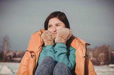 Woman looking away sitting with hands on chin against sky during winter