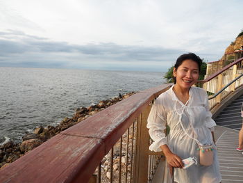 Portrait of smiling young woman standing by railing against sea