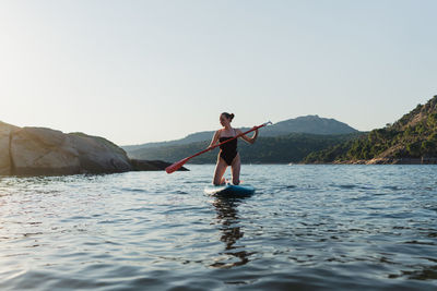 Full body of active female in swimwear floating on paddleboard in rippling lake against rock and shore with forest on summer day