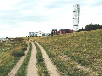 Dirt road amidst grassy field by turning torso in city against sky