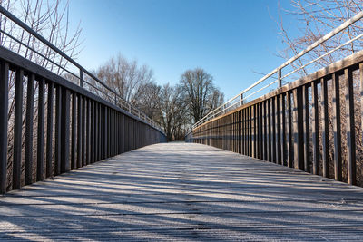 Narrow footbridge along bare trees against clear sky