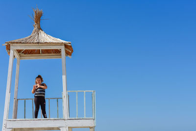 Girl standing in lifeguard hut while making heart shape gesture against sky