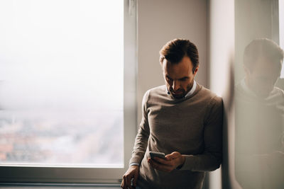 Young man using phone while standing on window