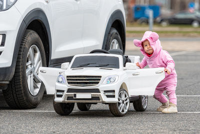 A little baby girl at a big white childrens electric toy car in the parking lot next to a real car