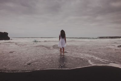 Rear view of woman standing at beach against cloudy sky