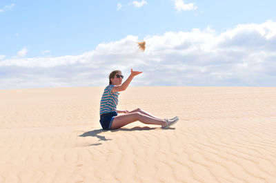 Full length side view of teenage girl throwing sand while sitting on desert against sky during sunny day