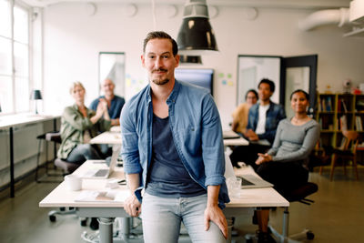 Portrait of confident businessman leaning on table while coworkers sitting in background at work place