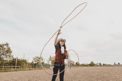 Cowgirl throwing lasso while standing at ranch against sky