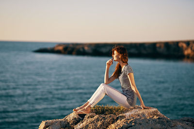 Man sitting on rock by sea against sky during sunset