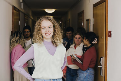 Portrait of smiling young woman with multiracial friends in background at corridor in college dorm