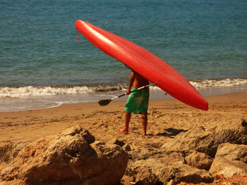 Rear view of man carrying red canoe on shore at beach