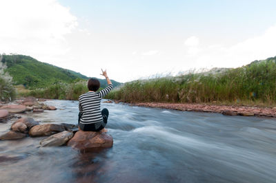 Rear view of woman gesturing horn sign while sitting on rock in river