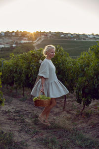 Woman with a wicker basket of green grapes stands in her vineyard at sunset