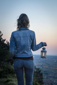 Rear view of woman holding lantern while standing on field against clear sky during sunset