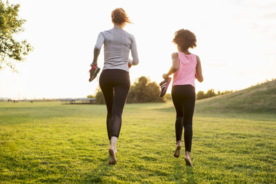 Rear view of woman and girl jogging while holding shoes on grass during sunset