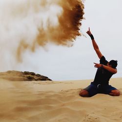 Full length of young man with hand raised kneeling on sand at desert against sky