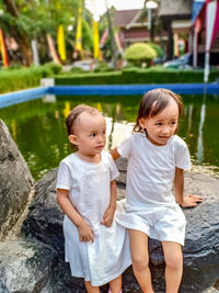 Cute siblings looking away while sitting on rock by pond in park