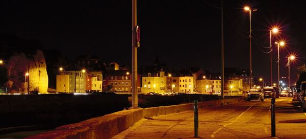 Illuminated street by buildings against sky at night