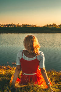 Rear view of woman sitting by lake against sky during sunset