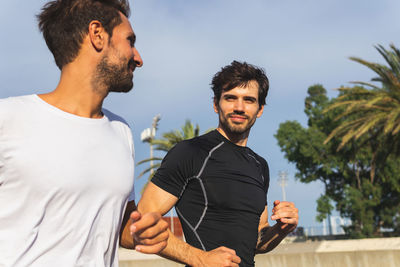 Hispanic male athletes looking at each other and jogging in park against cloudy blue sky and sea during cardio workout on sunny day