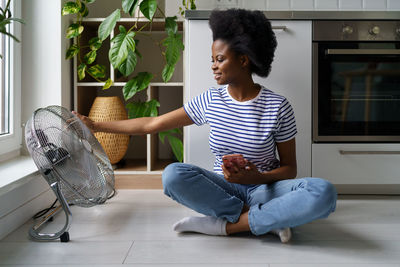 Satisfied african american woman enjoying cool air coming from behind electric fan and holds phone