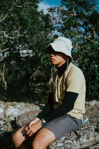 Young man wearing hat sitting on rock sitting against tree