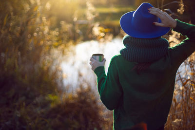 Woman in knitted sweater and blue hat stands bushes sunset and drinks tea from an iron thermos mug
